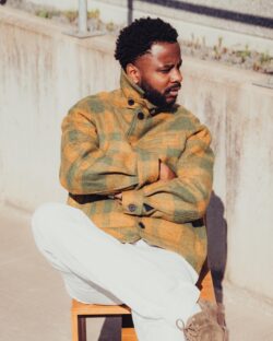 Hiram Bonheur Tamundele sits on a wooden chair outdoors, wearing a green and orange plaid jacket, white pants, and brown shoes, with his arms crossed and looking to the side.