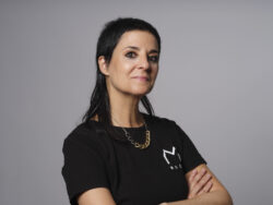A woman with a black T-shirt and a wolfcut looks at the camera, her arms are crossed