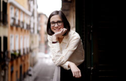 A woman rests her elbow on a balustrade and looks at the camera smiling; she has mid-length hair and wears glasses