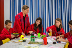 Four children in red craft clothes with paint stains are busy painting together with their supervisor.