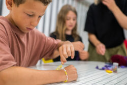 Een kind draagt een vriendschapsarmband terwijl het eraan werkt. Op de achtergrond is een tweede kind te zien dat met hulp van de begeleider aan het knutselen is.