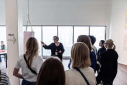 A museum guide presents to an audience, highlighting an exhibit with her finger while explaining its significance.