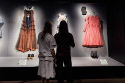 Two young people admire dresses on display in a museum, engaged in conversation about the fashion exhibits.