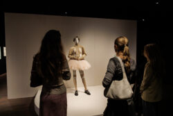 Drie jongeren bewonderen een mannequin met een roze tutu in een tentoonstelling.