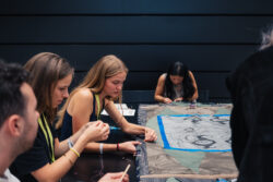 Several people sit at a table, focused on sewing or embroidering fabric together in a well-lit indoor space.