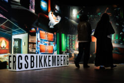 Two people stand in a dimly lit room, observing brightly colored wall art and a large sign reading "DIRK BIKKEMBERGS." The atmosphere is immersive and reflective.
