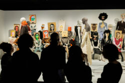 A group of people views mannequins dressed in various outfits, some with colorful, abstract faces drawn on paper or cardboard covering their heads.