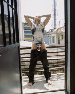 A man in low-sacked pants and a tank top stands on a balcony, holding his head with eyes closed, embodying Diesel's pride campaign.