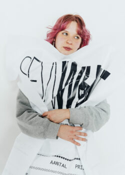 A person with pink hair and a gray sweatshirt holds a large white sheet with black printed text, standing against a white background.