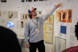 A young MoMu guide in a cap points to a timeline wall of newspaper clippings, sketches and photographs around the year 1980.