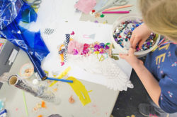 Child embellishes a female silhouette on paper with various types of textile and small sequin to achieve a 3D design.