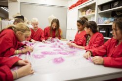 Des enfants vêtus de tabliers rouges sont assis autour d'une table pour étudier un grand échantillon de tissu.