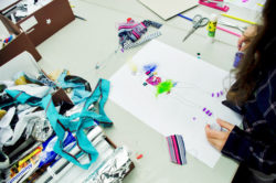 Child embellishes a female silhouette on paper with various types of textile and small sequin to achieve a 3D design.