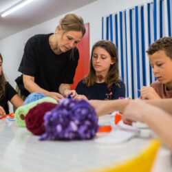 Four children listen attentively to the supervisor at a table full of colorful craft supplies.