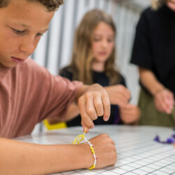 Een kind draagt een vriendschapsarmband terwijl het eraan werkt. Op de achtergrond is een tweede kind te zien dat met hulp van de begeleider aan het knutselen is.