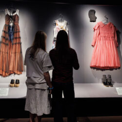 Two young people admire dresses on display in a museum, engaged in conversation about the fashion exhibits.