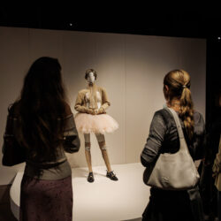 Drie jongeren bewonderen een mannequin met een roze tutu in een tentoonstelling.