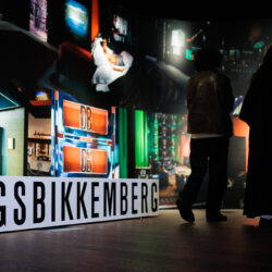 Twee mensen staan in een schemerige ruimte en kijken naar felgekleurde wandkunst en een groot bord met de tekst “DIRK BIKKEMBERGS”. De sfeer is meeslepend en contemplatief.