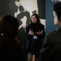 Een vrouwelijk gids geeft uitleg aan een groep mensen voor een zwart-wit afbeelding van Dries van Noten