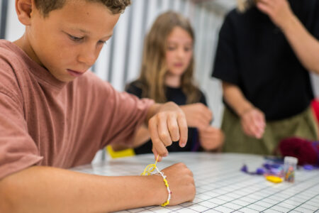 Een kind draagt een vriendschapsarmband terwijl het eraan werkt. Op de achtergrond is een tweede kind te zien dat met hulp van de begeleider aan het knutselen is.