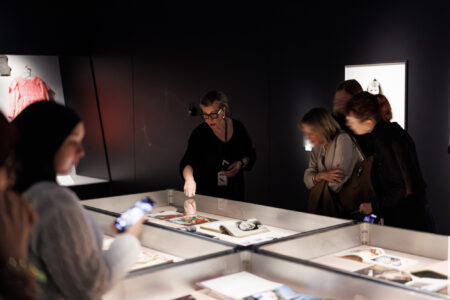 Een gids wijst naar voorwerpen achter glas terwijl bezoekers de tentoonstelling bekijken en discussiëren over de tentoongestelde artefacten.