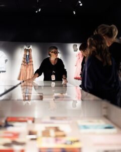 Een museumgids, een vrouw, staat voor een vitrine met voorwerpen en boeit bezoekers met haar kennis.