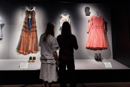 Twee jongeren bewonderen jurken die in een museum worden tentoongesteld en voeren een gesprek over de mode-expositie.