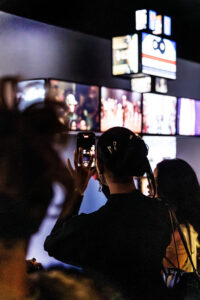 A group of people looks at an installation by Walter Van Beirendonck, while one person takes a photo of television screens shaped like a robot.