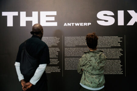 Two people stand reading information on a wall about "The Antwerp Six." The text is bold and white on a black background, creating a reflective mood.