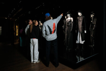 A young guide points at Ann Demeulemeester mannequins in black leather and tailored clothing in a dark gallery during a Young Voices tour.