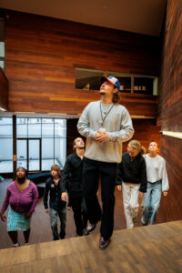 A young guide and a group of visitors look upwards as they climb a staircase in a museum hall with wooden walls.