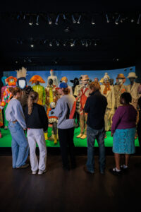 Young people seen from behind look at a Walter Van Beirendonck display of colourful mannequins in bold outfits and hats, set against a green floor and blue backdrop