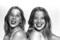 Two young girls pose together, both sticking out their tongues; one girl is smiling joyfully at the camera.