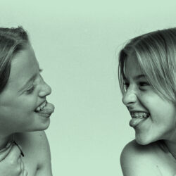 Two young girls are laughing and smiling while playfully sticking out their tongues at each other.