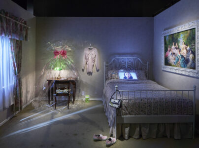 A vintage bedroom features a floral twin bed with a television on the pillow. Nearby, a draped pink dress and framed art add a whimsical, nostalgic touch