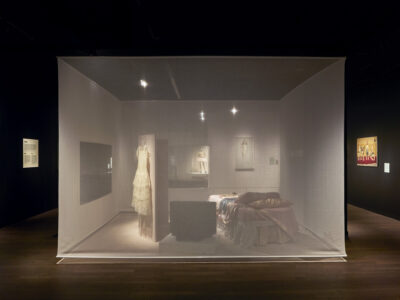 A softly lit museum exhibit displays a vintage bedroom scene behind a translucent curtain. The cozy room features elegant clothing and art, evoking nostalgia