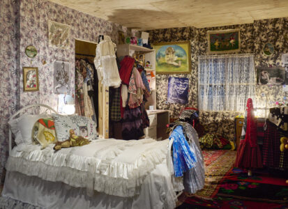 A cozy bedroom with floral patterned wallpaper, a neatly made bed with white ruffled linens, and vibrant, colorful clothing hanging on a rack