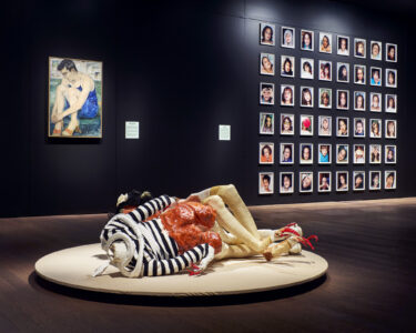 Exhibition space with a modern sculpture on a round platform, a painting of a seated figure on the wall, and a grid of diverse portraits displayed