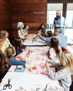 A group of parents and children are engaged in creative activities at a colorful table in the hall of a museum.