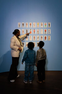 Een vrouw wijst modesilhouetten aan die gedrukt zijn op een blauwe muur, twee jonge kinderen kijken met interesse.