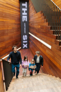 Twee jonge ouders en hun twee dochtertjes lopen de trap op, al lezend, met op de achtergrond het spandoek voor de tentoonstelling ‘The Antwerp Six’.
