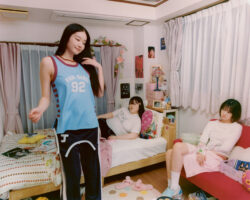 Three young women in a cozy bedroom. One stands wearing a "Fan Girl 92" shirt, another lounges on a bed, and a third sits on a red couch. The room is decorated with pink accents, flowers, and posters, conveying a relaxed and friendly atmosphere.