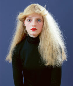 Young person with voluminous blonde hair in a black turtleneck against a blue background