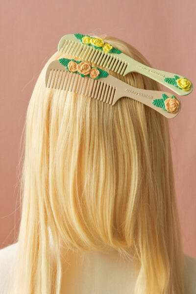 A mannequin bust with long blonde wig features two beige combs adorned with yellow and peach floral designs, set against a soft pink background