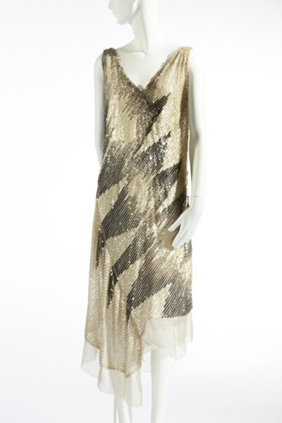 Evening dress in beige silk muslin covered with transparent grey and white sequins in a diagonal stripe pattern. Straight, loose fit, reaching approximately calf height, sleeveless. V-neckline at the front and back