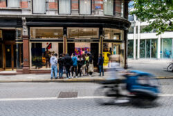 A group of young people listens to a guide on the corner of a street, while a cyclist passes by.