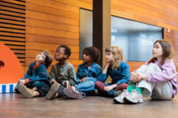 Een aantal kinderen zit op de grond en speelt met een groot oranje speelgoed, omringd door een speelse sfeer.