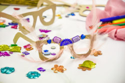 Kartonnen bril gemaakt door kinderen tijdens de Olivier Theyskens expo, gedecoreerd met rose veren en paarse en blauwe glitters en steentjes.
